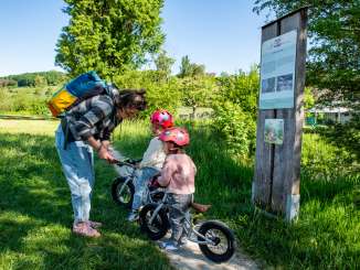 Biberweg Thayngen Eine erwachsene Frau mit zwei Kindern auf einem Pfad neben einem Fluss. Beide Kinder sitzen auf einem Laufrad. Daneben befinden sich eine Tafel, wo ein Bild eines Bibers zu sehen ist.