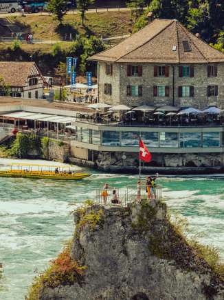 Mittelfelsen Rheinfall Auf dem Felsen mitten im Wasserfall stehen mehrere Personen und eine Schweiz Flagge. Hinter dem Felsen sieht man aufs Schlössli Wörth.