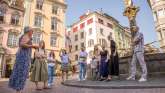 Eine Gruppe von Personen in einer Altstadt neben einem Brunnen hören einer Person gespannt zu.
