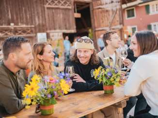 Eine Gruppe an Freunden sitzt an einem Festbank, welcher mit Blumen geschmückt ist. Im Hintergrund ist eine Scheune zu sehen. Die Freunde stossen mit einem Glas Wein an.