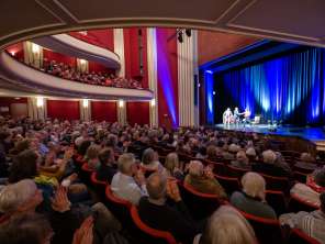 Ein Sall gefüllt mit Menschen, welche mit den Händen klatschen. Eine beleuchtete Bühne mit drei Personen darauf sowie ein Sofa und ein Sessel.