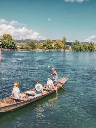 Weidlingsfahrt auf dem Rhein Drei Personen und ein Hund sitzen im Weidling. Das Holzboot treibt gemütlich den Rhein hinunter. Eine Frau steht, hat ein Ruder in der Hand und steuert das Boot.