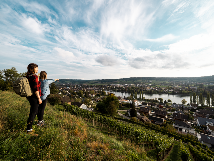 Zwei Wanderer stehen auf einem Feld oberhalb der Reben und der Stadt Stein am Rhein und geniessen die Aussicht auf die Altstadt und den Rhein.