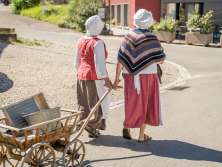 Zwei Wöschwiiber laufen mit einem Leiterwagen aus Holz durchs Dorf. Sie tragen eine weisse Haube.