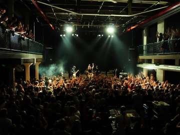 Eine Menschen gefüllte Halle. Links und rechts hat es ein Balkon. Vorne befindet sich eine Bühne mit einer Band, welche ein Konzert spielt.