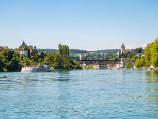 Blick vom Rhein auf den Munot und eine Eisenbahnbrücke. Richtung Schaffhausen fährt das Kursschiff.