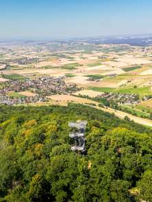 Siblinger Randenturm mit Dorf Im Vordergrund sieht man einen Turm, der aus dem Wald ragt. Im Hintergrund sieht man eine flache Landschaft mit vielen Dörfern und Feldern.