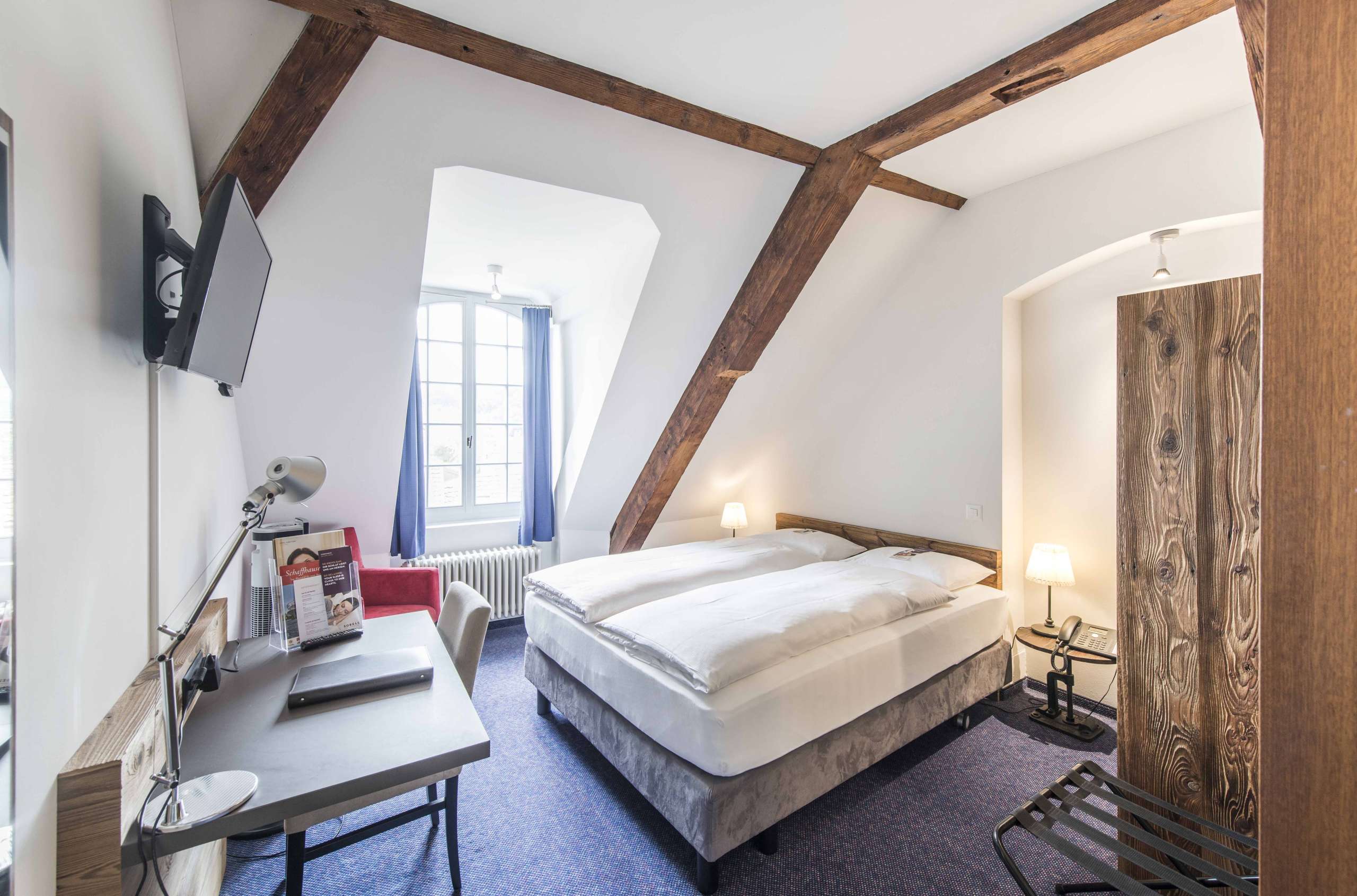 Hotelzimmer im Sorell Hotel Rüden Schaffhausen mit einem Doppelbett und Nachttischen, einem Schreibtisch, einem Schrank, einem Fernseher und einem roten Sessel.