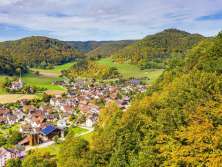 Blick vom Wald auf ein Dorf umgeben von herbstlichen, waldbedeckten Hügeln. Leicht ausserhalb des Dorfes steht eine Kirche.