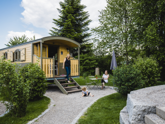 Ein Zirkuswagen aus Holz mit einem Kiesvorplatz. Es sind zwei Erwachsene Personen zu sehen. Eine davon läuft dir Treppe hinunter vom Zirkuswagen. Eine weiter Person sitzt auf einem Stuhl auf dem Vorplatz. Ein weiteres Kind spiel am Boden mit dem Kies.