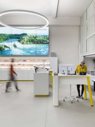 Visitor Centre Schaffhausen Das ist der Schalterbereich des Visitor Centres Schaffhausen. An einem Stehtisch steht eine Frau in senfgelben Pullover und telefoniert. Eine weitere Person läuft zum freien Schalter.