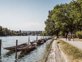 Vier Wanderer laufen am Ufer des Rheins entlangs. Im Rhein befinden sich diverse Weidlinge. In der Ferne ist die Festung Munot sowie die Stadt Schaffhausen zu sehen.