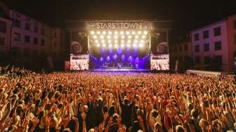 Stars in der Stadt Viele Personen mit den Armen in der Luft sind an einem Konzert mit einer Band auf einer grossen Bühne.