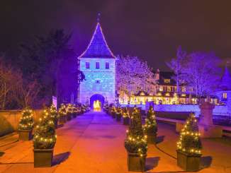 Ein Schloss ist in blauem/violetten Licht beleuchtet. Vor dem Eingang stehen kleine Weihnachtsbäume mit Lichterketten.