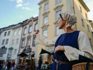 Die als Krämerin verkleidete Person zeigt mit ihrem Arm auf eine Sehenswürdigkeit.