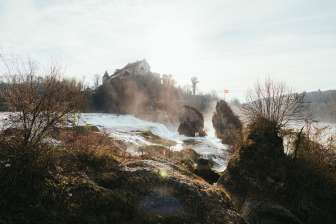 Ein Wasserfall mit einem Mittelfelsen. Auf dem Felsen ist eine Schweizer Fahne. Im Hintergrund ist ein Mittelalterliches Gebäude.
