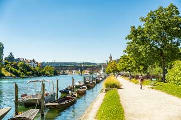 Flaniermeilie direkt am Rhein. Am Ufer stehen diverse Holzboote im Wasser und im Hintergrund sieht man die Festung Munot.