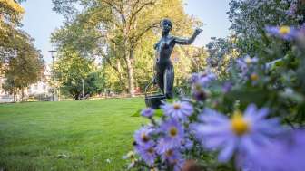 Eine Frauenstatue welche einen Korb trägt in einem Park. Im Vordergrund hat es blühende Blumen und im Hintergrund hat es Bäume.