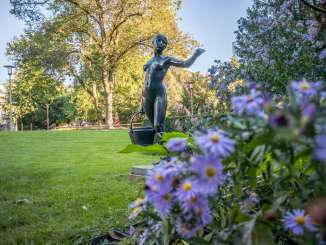 Eine eiserne Skulptur steht auf einer Wiese. Sie bildet eine Frau ab, die einen Eimer Wasser trägt.